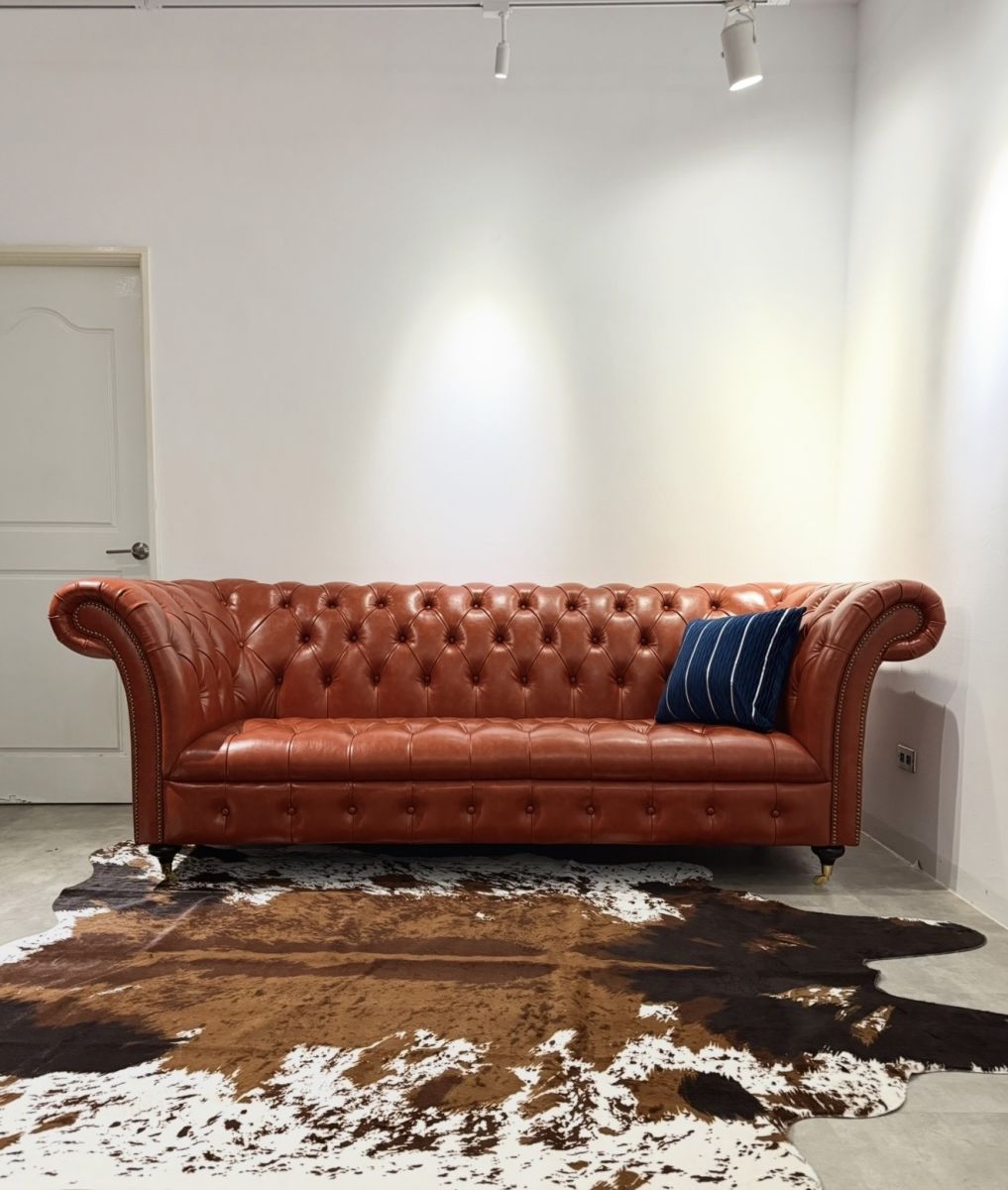 Chesterfield Wanda系列焦糖色仿真PU皮沙發側邊特寫，展現經典菱格紋拉扣工藝與復古黃銅滾輪，搭配牛皮地毯與藍色條紋抱枕。