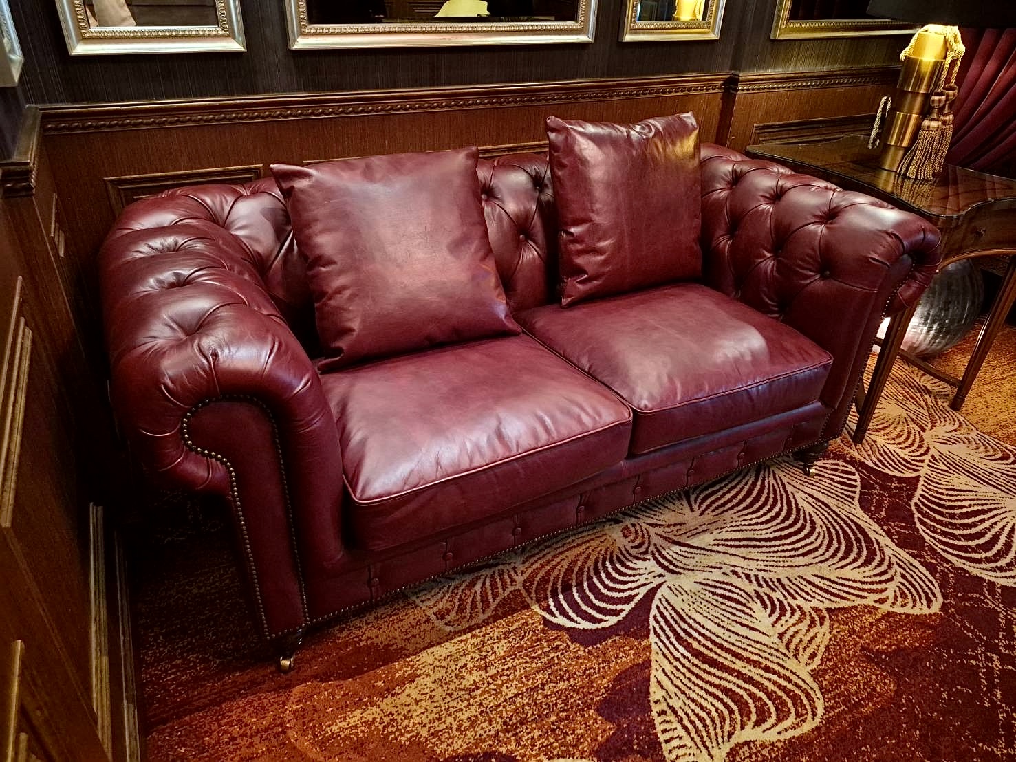 近距離展示 Chesterfield 酒紅色牛皮沙發的半苯染工藝皮革質感、深扣拉花與邊緣整齊的黃銅鉚釘。